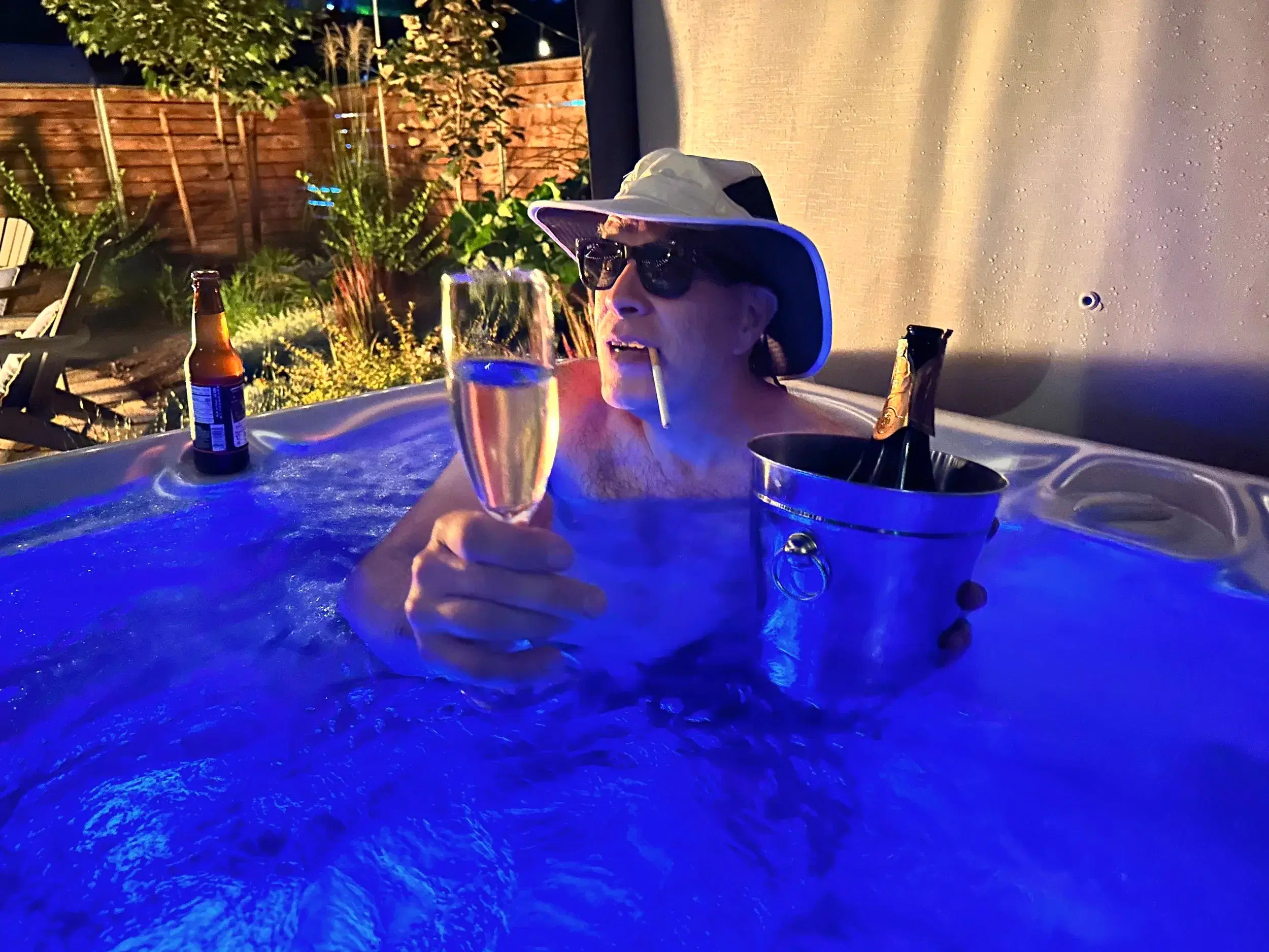 A guest in a hot tub at night, lit blue, raising a glass of champagne.