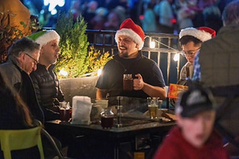 Friends in Santa hats sharing drinks on a lit patio on Parade of Lights night.
