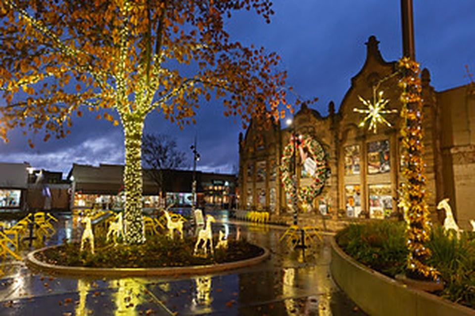 Lit reindeer figures and a light-wrapped tree in a downtown plaza on a rainy winter evening.