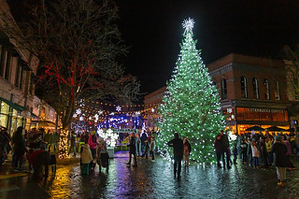 A crowd gathered around a tall lit Christmas tree on a wet downtown Walla Walla street at night.