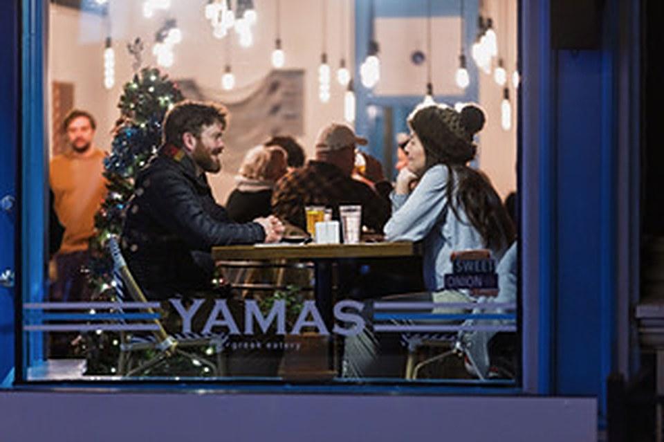 A couple sharing drinks at a window table inside Yamas, a Christmas tree glowing behind them.