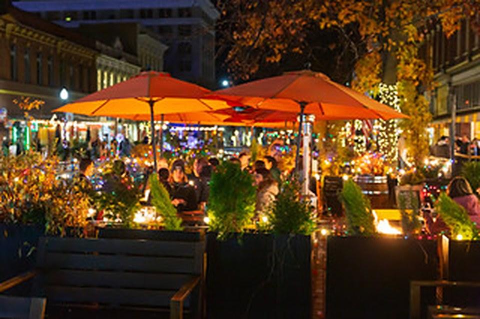 Diners under orange umbrellas on a downtown patio strung with lights at night.